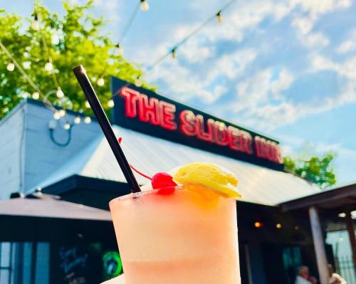 A refreshing cocktail with a cherry and lemon slice, in front of a restaurant sign that reads "The Slider Inn."