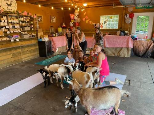 A group of children and adults sit on mats surrounded by playful goats in a cozy barn setting.