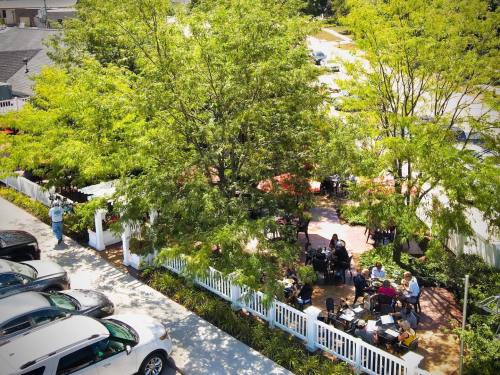 Aerial view of a patio surrounded by trees, with people dining at tables and parked cars nearby.