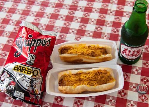 Two chili cheese hot dogs topped with shredded cheese, a bag of BBQ potato chips, and a green soda on a checkered tablecloth.
