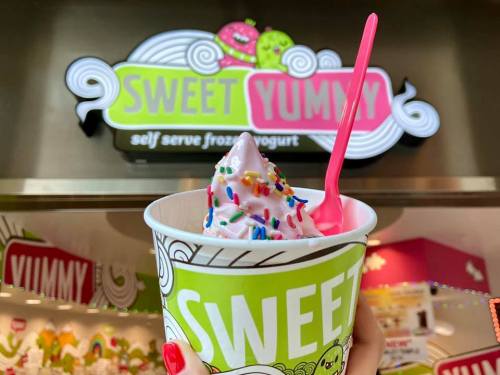 A hand holds a cup of pink froyo topped with sprinkles in front of a "Sweet Yummy" self-serve frozen yogurt sign.