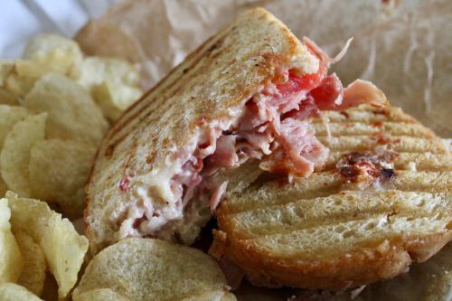 A grilled sandwich with ham and cheese, partially eaten, served with a side of potato chips.