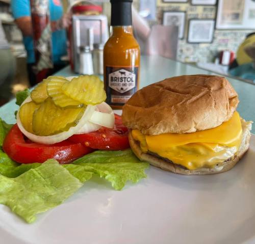 A cheeseburger with lettuce, tomato, pickles, and a bottle of Bristol hot sauce on a diner table.