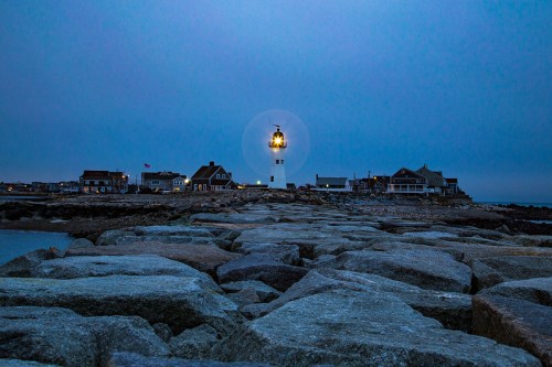 A lighthouse shines brightly at dusk, surrounded by rocky shores and quaint buildings under a cloudy sky.