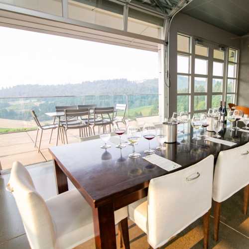 A dining table set with wine glasses overlooks a scenic view of hills and greenery through large windows.