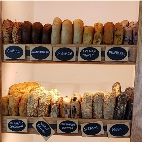 A display of various bagels labeled with flavors like garlic, spinach, and blueberry on wooden shelves.