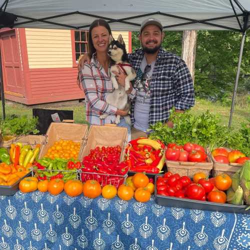 A couple holds a dog at a farmers market, surrounded by colorful fresh vegetables and fruits on a table.