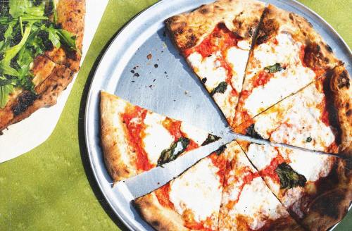 A pizza with melted cheese and basil, sliced into pieces, on a silver tray, next to a salad on a white plate.