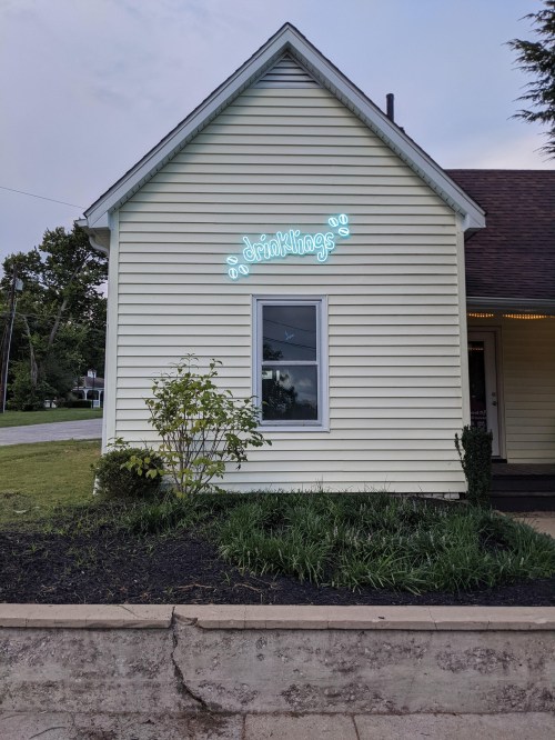 A light blue neon sign reading "drinklings" on the side of a yellow house, with greenery in front.