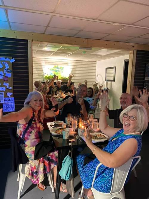 A group of people at a table celebrating, smiling, and raising their hands in a lively restaurant setting.