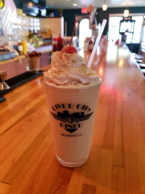 A tall milkshake topped with whipped cream and a cherry, served in a glass with "River City Diner" logo.