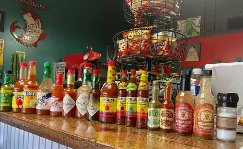 A variety of hot sauce bottles lined up on a wooden counter in a restaurant, with snacks in the background.