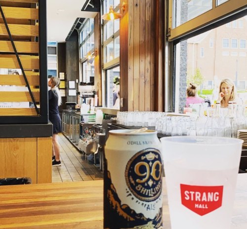 A bar interior with wooden accents, featuring a can of beer and a cup on the counter, and patrons in the background.
