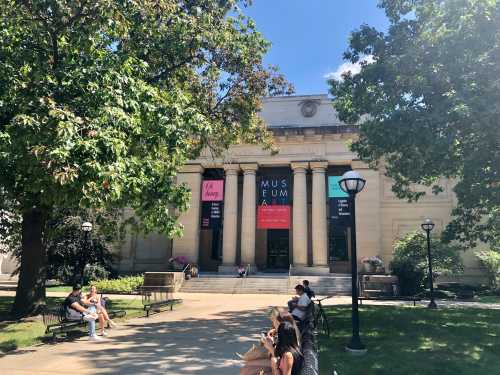 A sunny day at an art museum with people sitting on benches and trees providing shade in the foreground.