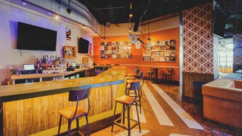 A modern bar interior featuring a wooden counter, empty stools, and colorful wall decor with shelves of bottles.