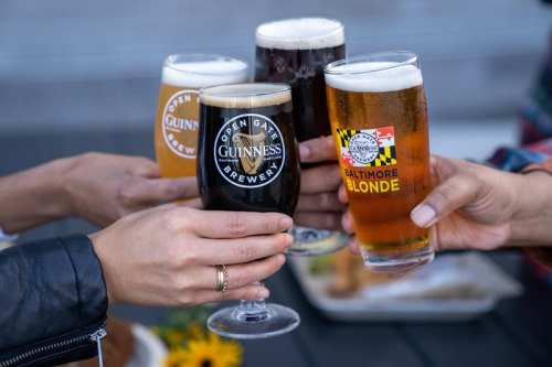 Four hands holding different beer glasses, including a dark stout and a light blonde, with a plate of food in the background.