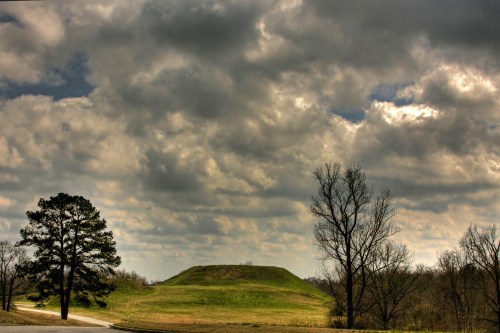 A grassy mound under a cloudy sky, with bare trees and a winding road in the foreground.