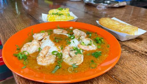 A vibrant orange plate with shrimp in sauce, rice, green onions, and sides of corn on the cob and bread.