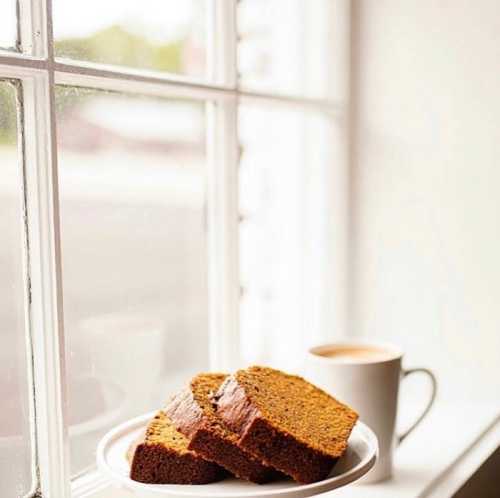 A plate with slices of cake sits on a windowsill next to a steaming cup of coffee. Natural light filters in.