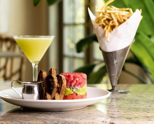 A plate with a colorful layered dish, a small pitcher, a cocktail, and a cone of fries on a table with greenery.