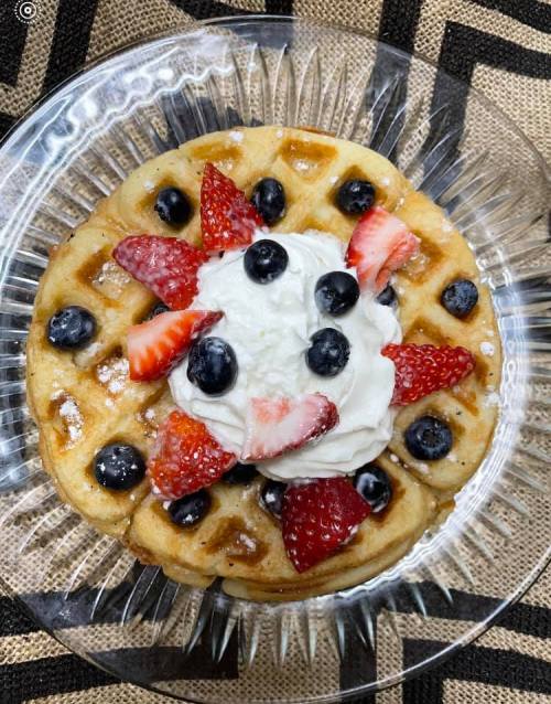 A waffle topped with whipped cream, strawberries, and blueberries, served on a clear glass plate.