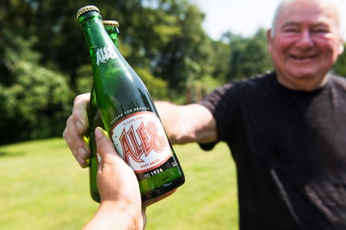 A person hands a green bottle of Ale-8 soft drink to an older man smiling in a sunny outdoor setting.