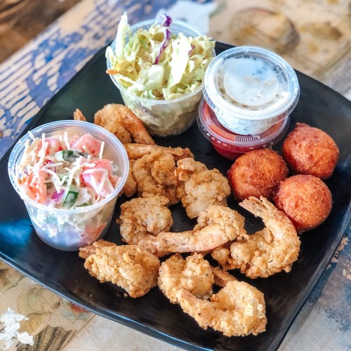A black plate with fried shrimp, coleslaw, two dipping sauces, and golden hushpuppies.