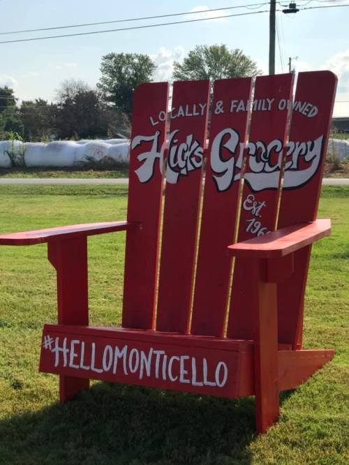 A large red Adirondack chair with "Hick's Grocery" painted on it, set in a grassy area with a hashtag below.
