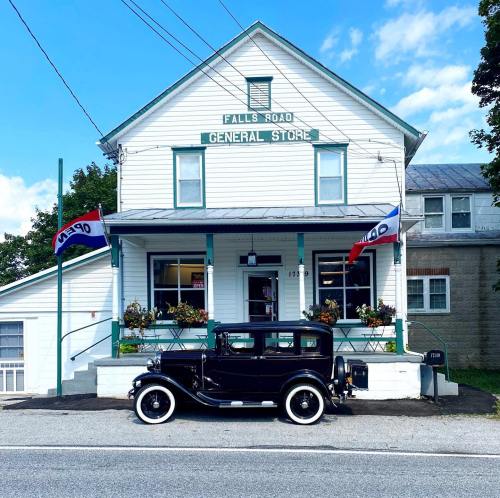 A vintage black car parked in front of a quaint general store with flags, surrounded by greenery and blue skies.