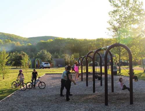 Children play on swings and bikes in a sunny park surrounded by trees and hills.