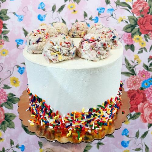 A round white cake topped with colorful sprinkles and cookie dough balls, set against a floral background.