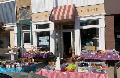 A storefront with a striped awning, featuring tables outside displaying various baked goods and snacks.