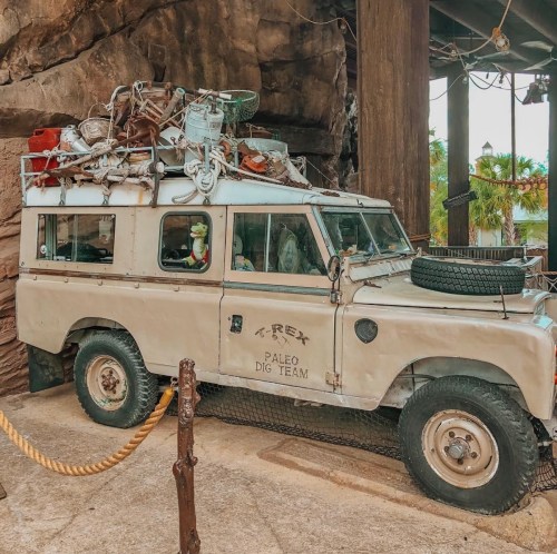 A vintage jeep with a cluttered roof rack, parked near a rocky structure, themed for a dinosaur dig site.
