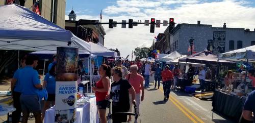 A bustling street fair with vendors, colorful balloons, and crowds enjoying the festivities under a blue sky.