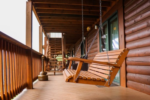 A wooden swing hangs on a porch of a log cabin, with stairs and railings visible in the background.