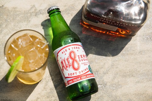 A green bottle of Ale-8-1 soft drink next to a glass of ice and a bottle of Woodford Reserve bourbon.