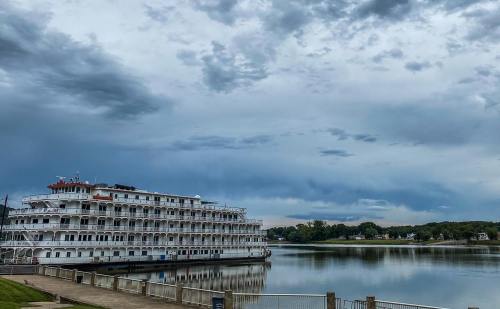 A large riverboat docked by the shore under a cloudy sky, reflecting in the calm water.