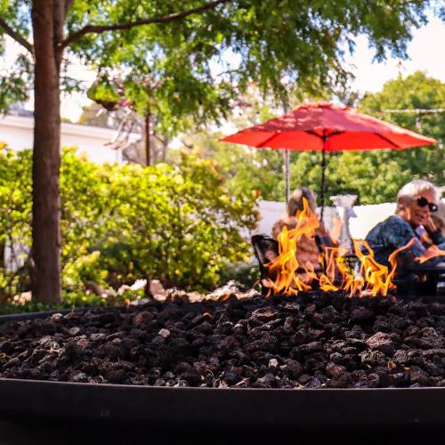 A cozy outdoor scene with a fire pit surrounded by greenery and people sitting under a red umbrella.
