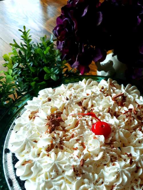 A delicious cake topped with whipped cream, chocolate shavings, and a cherry, surrounded by greenery and flowers.