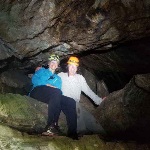 Two people in helmets pose inside a cave, surrounded by rocky formations and dim lighting.
