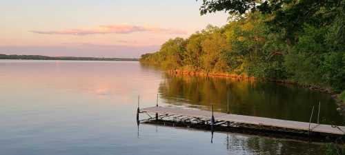 A serene lake at sunset, with a wooden dock extending into calm waters and lush greenery along the shore.