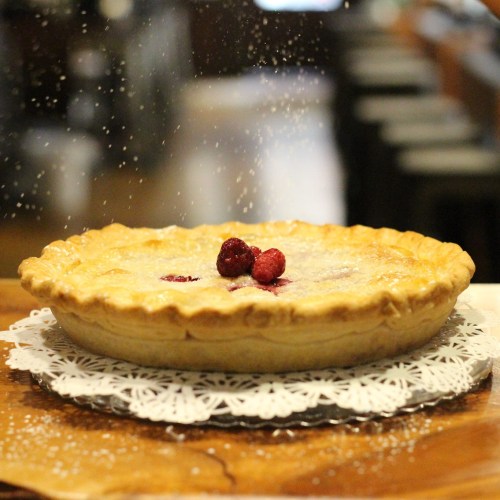 A freshly baked pie with raspberries on top, dusted with powdered sugar, on a decorative lace doily.