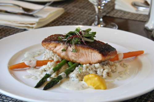 A plated dish featuring salmon on a bed of grains, garnished with asparagus, carrots, and a creamy sauce.