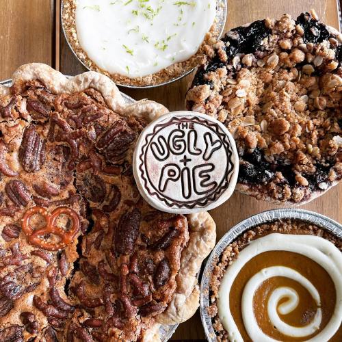 A variety of pies on a wooden table, including pecan, apple crumble, and a pie labeled "The Ugly Pie."