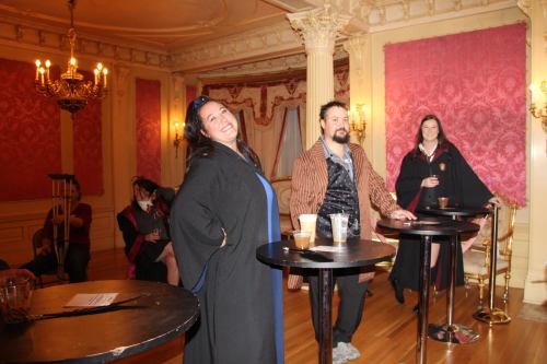 A group of people in costumes stands around tables in a lavishly decorated room, smiling and enjoying the event.