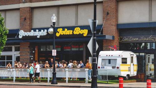 A bustling outdoor scene in front of a restaurant named "Goodnight John Boy," with a vintage camper parked nearby.