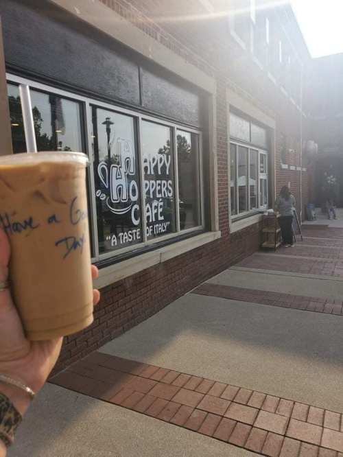 A hand holding an iced coffee in front of a café with a sign that reads "Happy Hoppers Café."