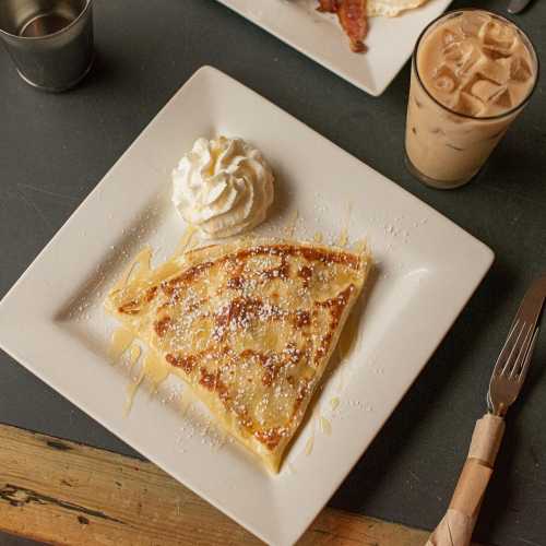 A plate with a folded crepe topped with whipped cream and powdered sugar, alongside a drink and a plate of bacon.