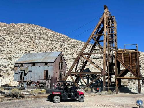 An old mining structure with a wooden frame and a nearby vehicle, set against a mountainous desert landscape.