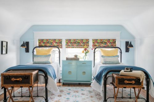 Brightly decorated bedroom with two metal beds, colorful window treatments, and a teal dresser between them.